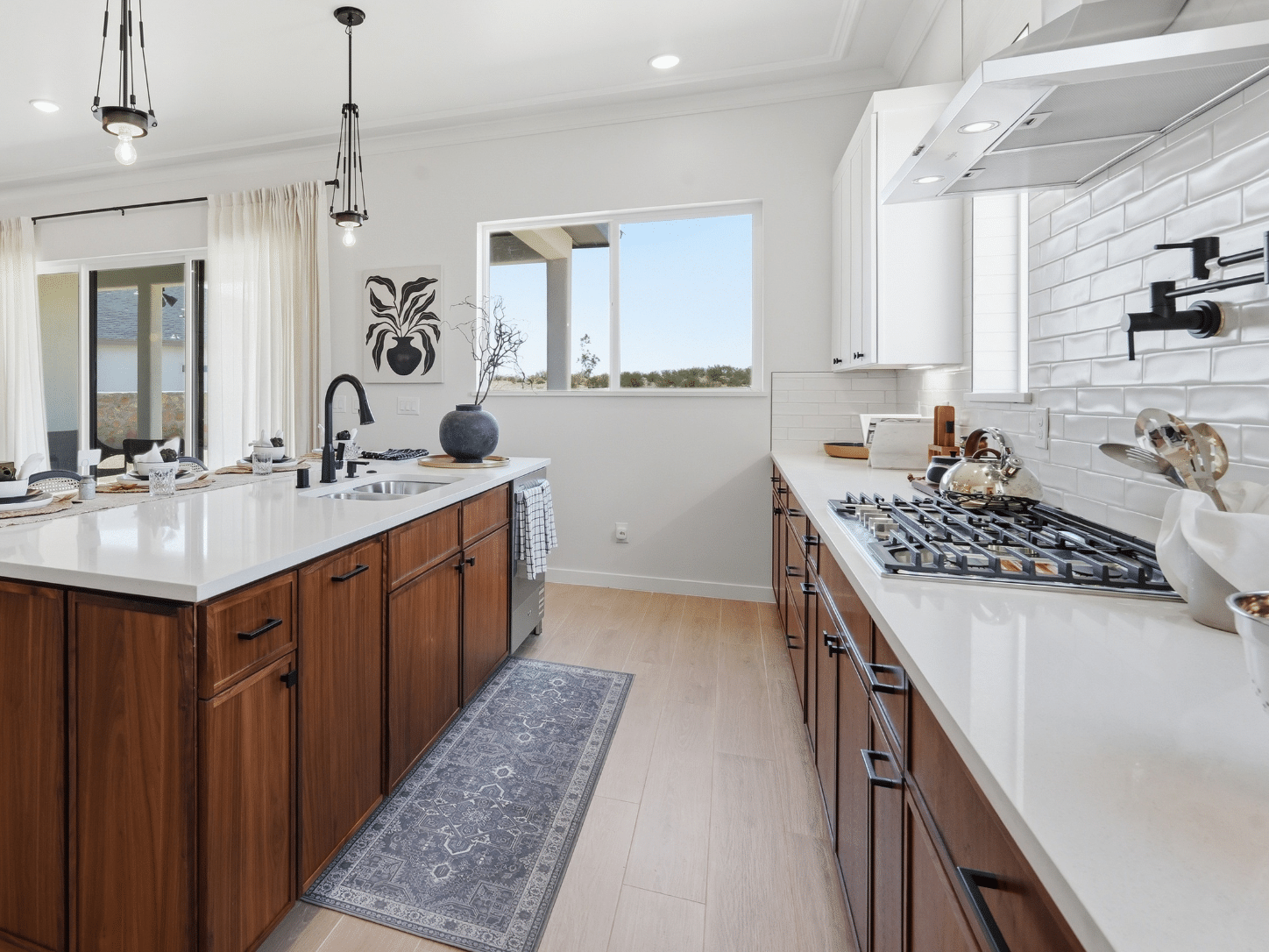 A sleek modern kitchen showcases wood cabinetry, crisp white countertops, and premium stainless steel appliances, including a gas stove. A central island with an integrated sink stands beneath expansive windows, bathing the space in natural light. Neutral decor complements light wood flooring, while elegant pendant lights complete the bright and welcoming atmosphere.