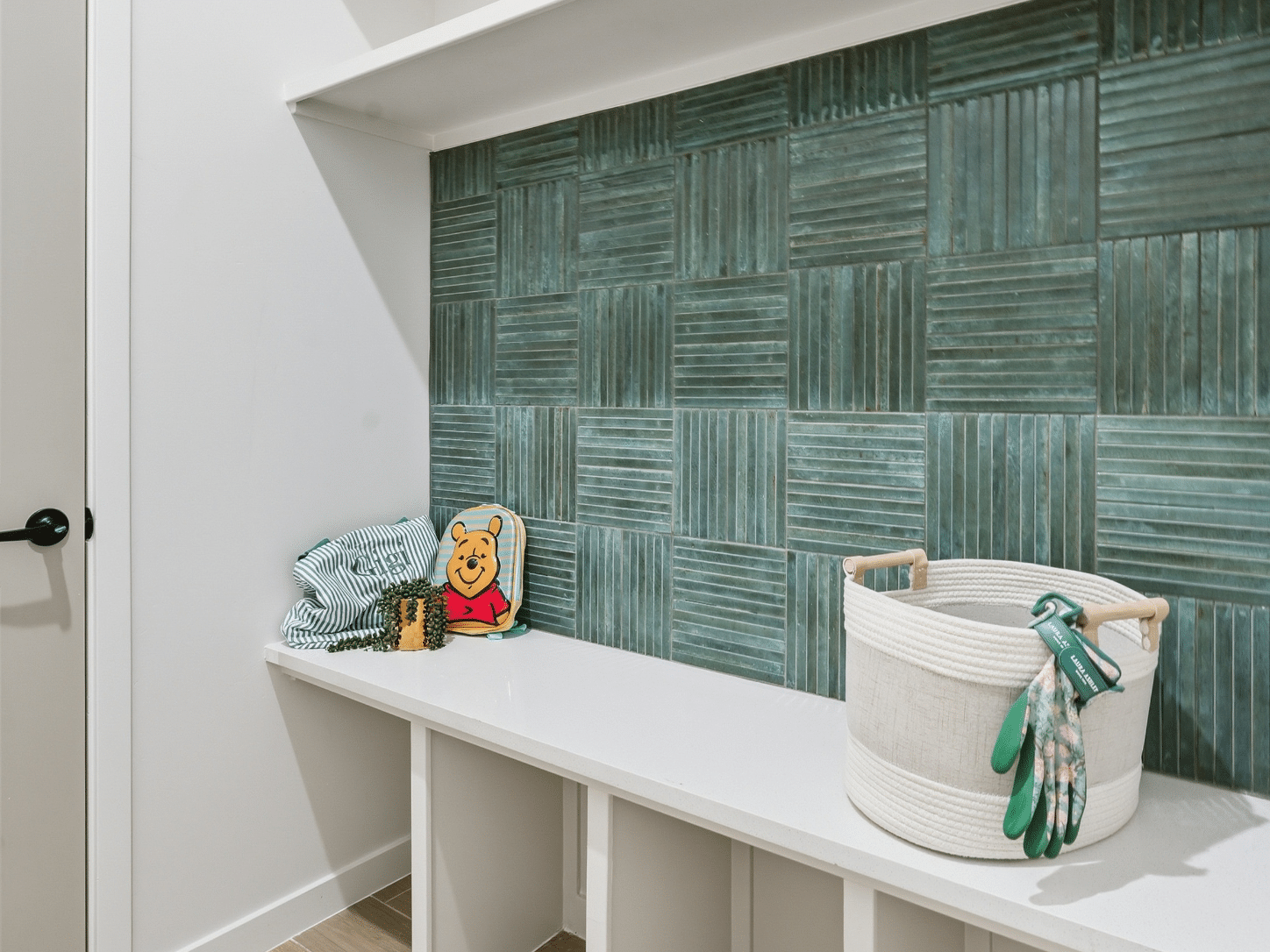 A well-appointed mudroom bench is adorned with a neatly folded striped towel and a whimsical Winnie the Pooh cushion. Beside them, a woven basket holds gardening gloves at the ready, set against a chic green textured tile backdrop.