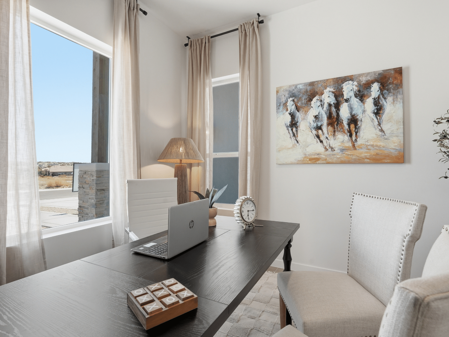 A sleek, contemporary home office features a black desk paired with crisp white chairs, complemented by a laptop, lamp, and clock. Natural light streams through the window, while a dynamic painting of galloping horses commands attention above the workspace. The room’s neutral palette fosters an atmosphere of tranquility and focus.