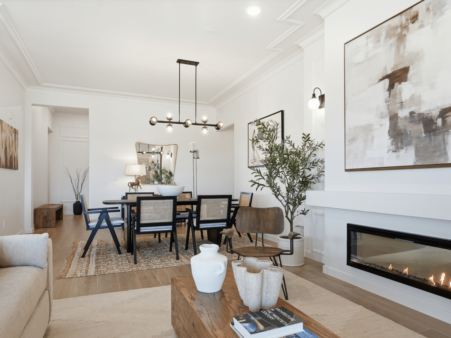 A sophisticated living and dining space is adorned in neutral hues, complemented by abstract artwork and sleek contemporary furnishings. A stylish fireplace anchors the room, while vibrant green plants and artful vases atop a wooden coffee table introduce warmth. Sunlight pours in, bathing the interior in crisp natural light.
