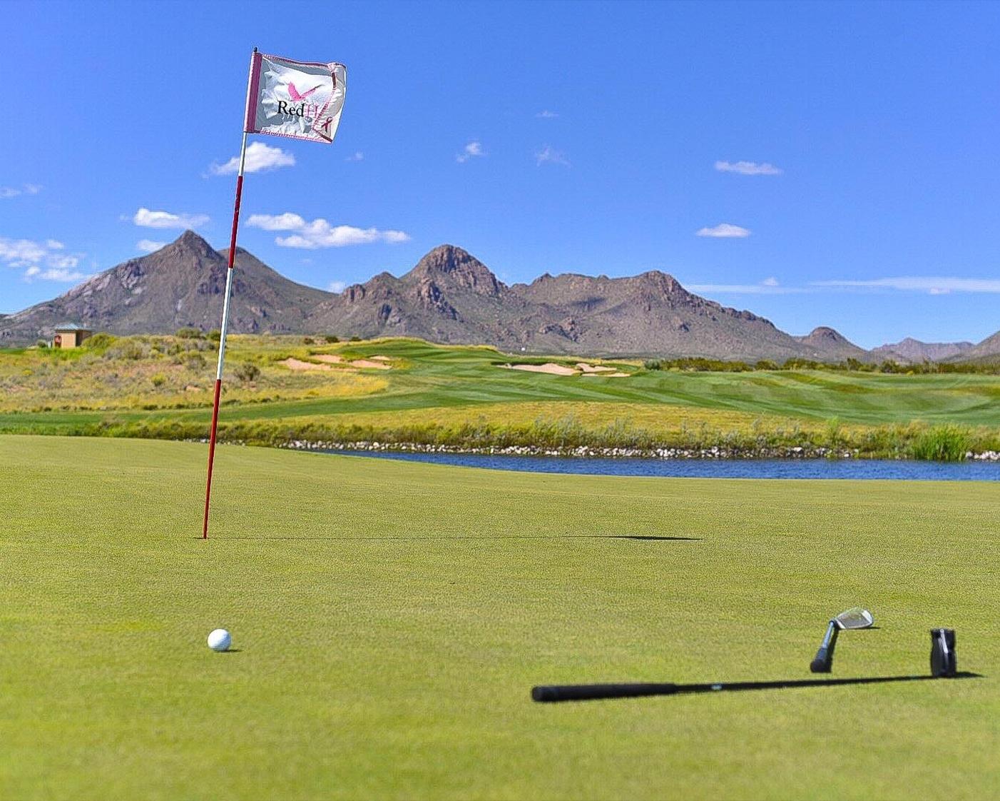 On a manicured golf course green, a flag marks the hole as a golf ball rests nearby, framed by a putter and club lying on the grass. Towering mountains rise in the distance beneath a clear blue sky.