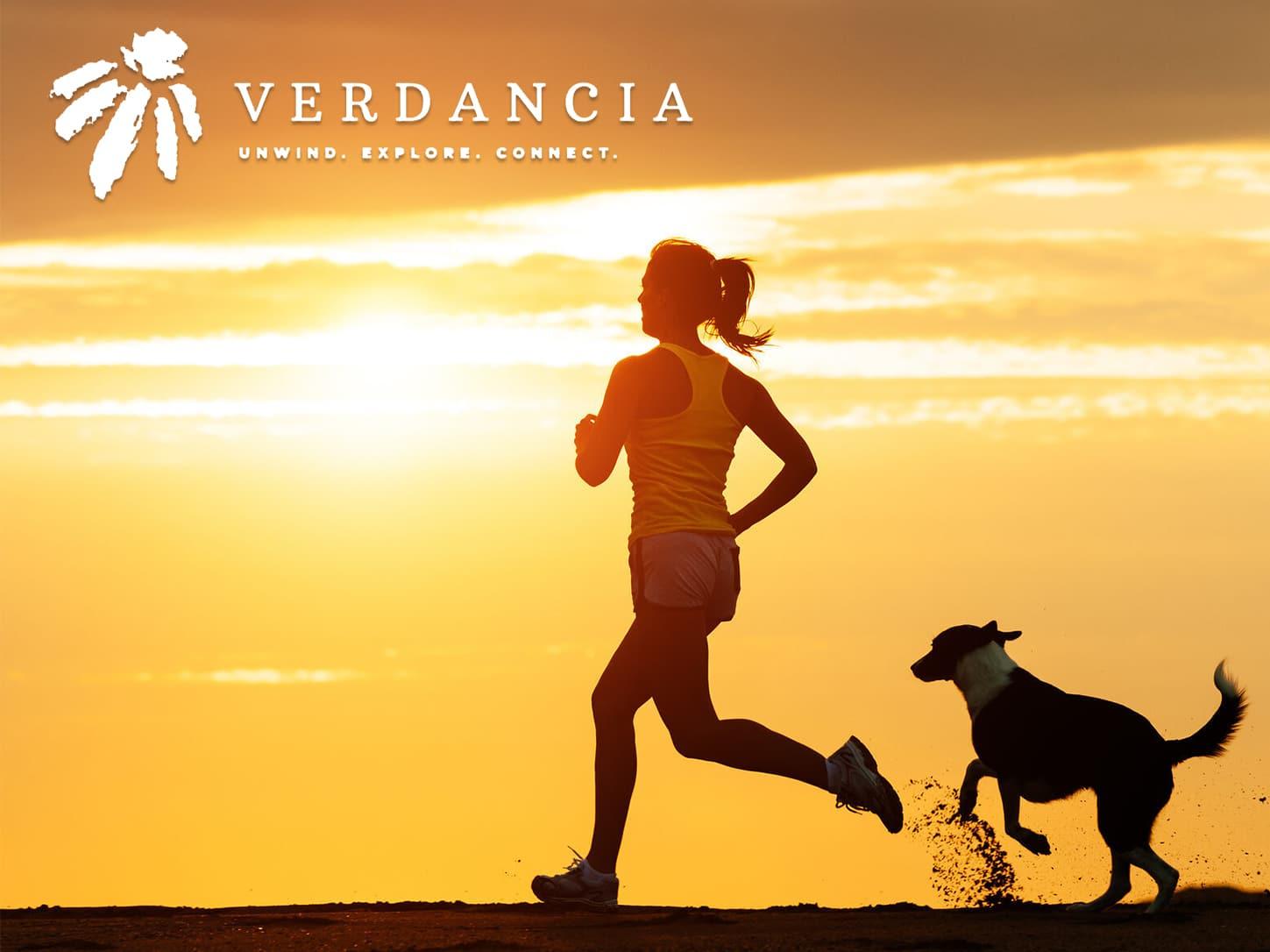 At sunset, a woman jogs beside her dog as the Verdancia logo and the slogan “Unwind. Explore. Connect.” appear prominently in the sky overhead.