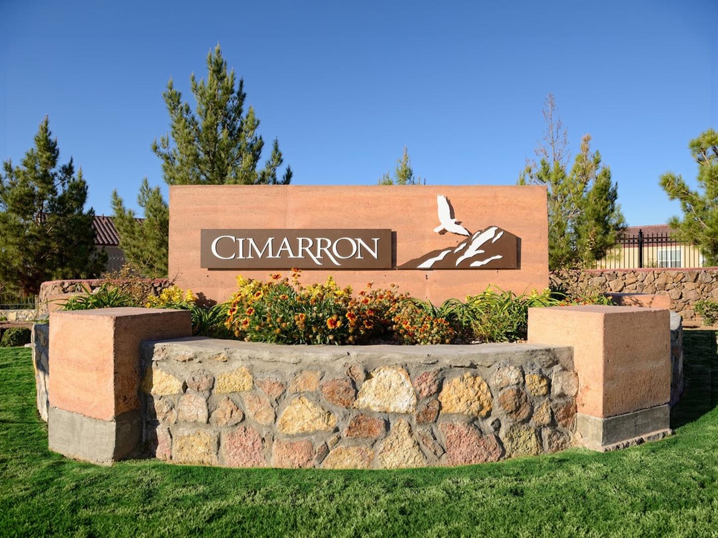 A stone sign bearing the inscription “CIMARRON” is adorned with a bird graphic perched above a mountain silhouette. Vibrant flowers and lush green shrubs frame the base, set against a backdrop of leafy trees and clear blue sky.